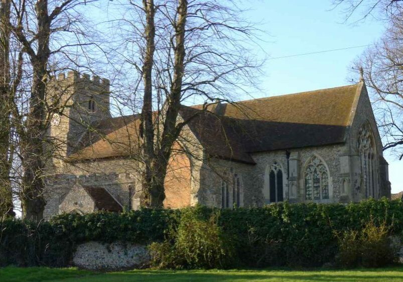 Exterior of a small church, seen through trees, a square tower, low, with clock.